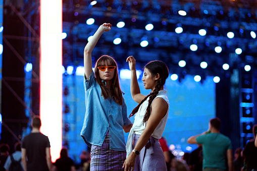 Two Asian and Caucasian girls dance in front of a stage and enjoy the atmosphere at a music festival.