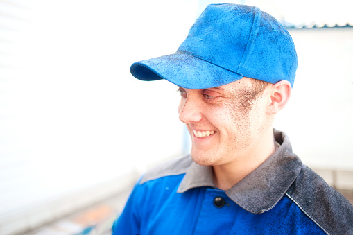 Portrait of young worker in work clothes and blue baseball cap with stained face. Cheerful and happy Caucasian engineer or locksmith smiles. Positive emotions after work done. Genuine guy.