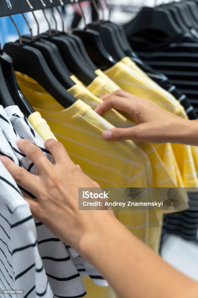 women's hands are sorting through T-shirts hanging on a hanger in the store. female hands sort through t-shirts hanging on a hanger in the store. A woman in a store looks through shirts hanging on hangers. woman's hands choose clothes. Shopping concept Collection Stock Photo