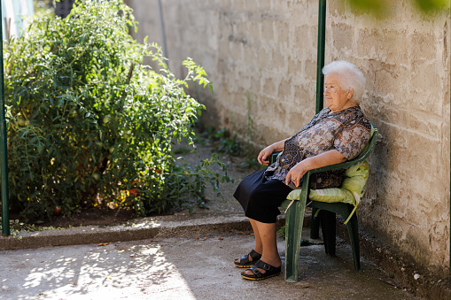 An old woman seats in a plastic chair in a backyard.