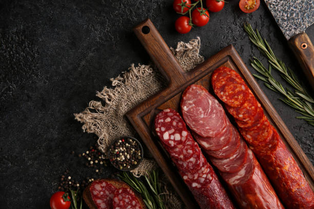 salsicha e frios. tipos de diferentes salsichas de pão em uma tábua de madeira velha no fundo preto e arroto com tomate cereja, alecrim, alho e especiarias. rustic.top. imagem de fundo, espaço de cópia - delicatessen meat cold cuts chopped - fotografias e filmes do acervo