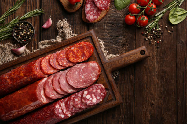 saucisse et charcuterie. types de différentes saucisses de pain sur un vieux fond en bois et toile de jute avec des tomates cerises, du romarin, de l’ail et des épices. rustique. retour au début. image d’arrière-plan, espace de copie - cold cuts thin portion salami photos et images de collection