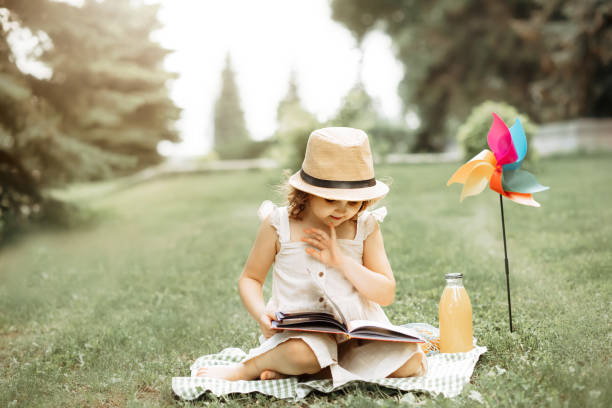 Cute little girl in a hat sitting on a blanket and reads a book at a picnic in nature stock photo