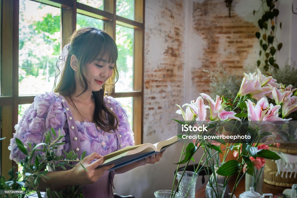 Girl in a princess dress reads a book in a room with windows and lots of green plants and lilies in a vase on the table like a fairy tale. Soft focus Adult Stock Photo