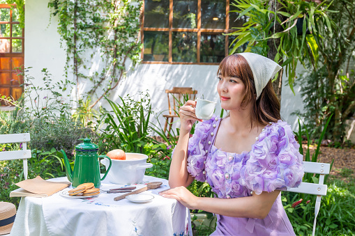 young woman in a princess dress sits drinking tea on a vintage table with cookies and books in a backyard with plants and flowers.