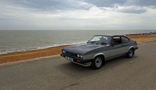 Felixstowe, Suffolk, England - May 01, 2022: Classic Grey and Silver Ford Capri being driven along seafront promenade beach and sea in background.