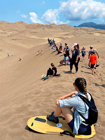 Alamosa County, Colorado, USA - July 11, 2021 - A woman sitting on a sandboard watches fellow tourists at Great Sand Dunes National Park enjoy sand-boarding on the dunes of the namesake recreation area.
