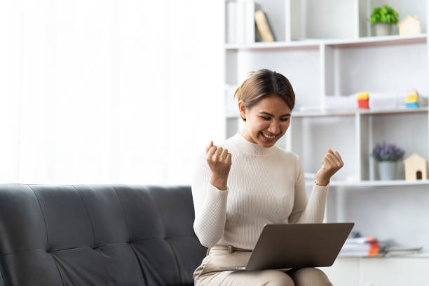 istock Young attractive asian female working at home hand use computer laptop checking business data chart planning report with leisure relax on sofa in living room home. 1417357789