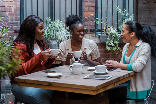 Medium shot of three female friends/co-workers having coffee/tea together on an outdoor terrace in the North East of England. They are laughing and talking together.