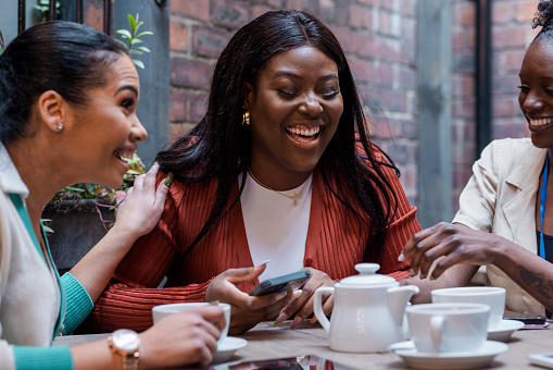 Medium shot of three female friends/co-workers having coffee/tea together on an outdoor terrace in the North East of England. They are laughing and talking together looking at a mobile phone.