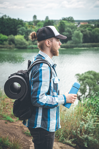 A male traveler with a backpack and a karemat holds a thermos in his hand, the concept of a hike.
