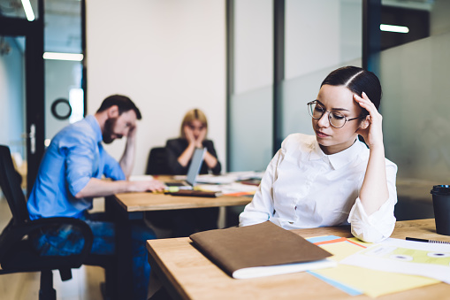 Weary young woman office worker in white formal shirt and glasses sitting over work and touching head with tired colleagues on background at coworking