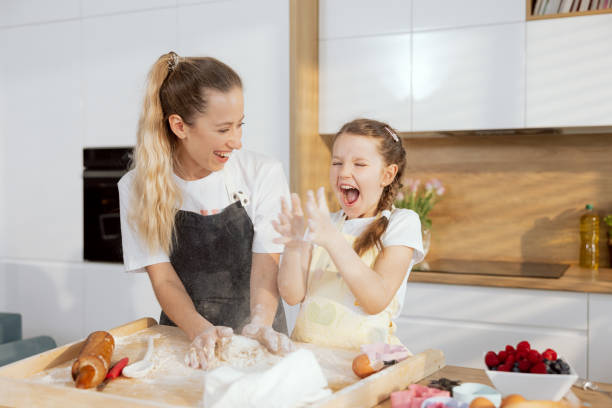 istock Beautiful older sister with younger one having fun in moder light kitchen cooking baking cake cookies biscuits. Kneading dough. Younger girl clapping in hands with flour 1417380592