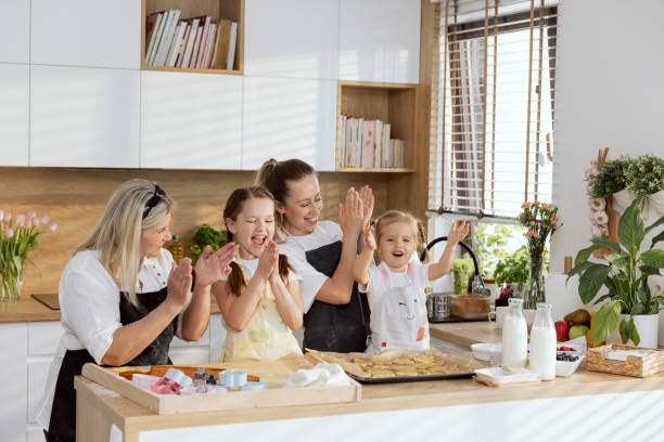 istock Delighted family wearing aprons clapping in hands smiling ending putting cookies biscuits on baking tray. Preparing dessert for breakfast standing at table in modern light beautiful kitchen 1417382846