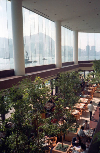 British Hong Kong, China - 1983: A vintage 1980's Fujifilm negative film scan of the downtown Hong Kong skyline across the water through large windows inside a restaurant, with tables and trees.