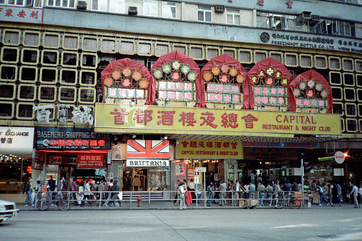 British Hong Kong, China - 1983: A vintage 1980's Fujifilm negative film scan of decorated ship signs and a crowd of pedestrians entering a building on the busy city streets in Hong Kong.