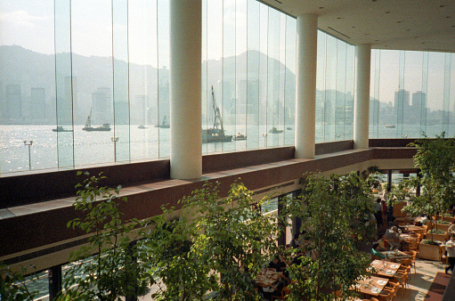 British Hong Kong, China - 1983: A vintage 1980's Fujifilm negative film scan of the downtown Hong Kong skyline across the water through large windows inside a restaurant, with tables and trees.