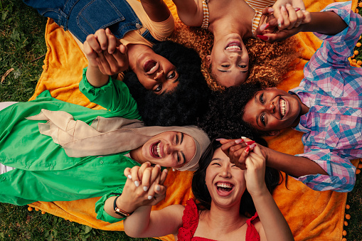 A group of joyful girls are lying in a circle on the lawn and holding their hands together.