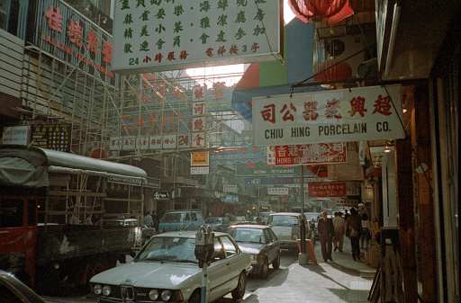 British Hong Kong, China - 1983: A vintage 1980's Fujifilm negative film scan of busy city streets covered in store signs in Hong Kong with pedestrians, cars, and shops.