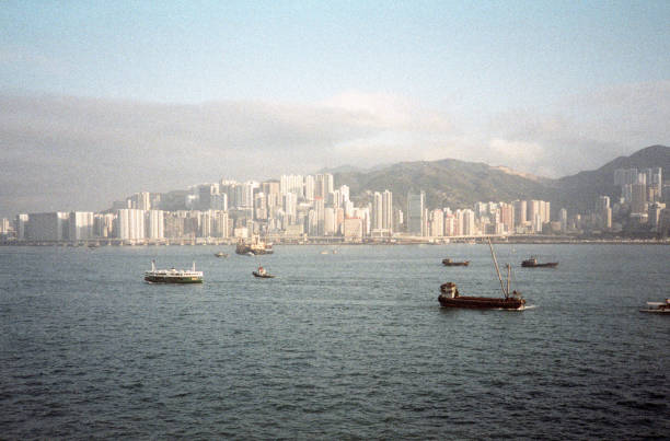 British Hong Kong, China - 1983: A vintage 1980's Fujifilm negative film scan of a photograph of the skyscrapers in downtown Hong Kong across the bay with multiple shipping container and fishing boats in the water.