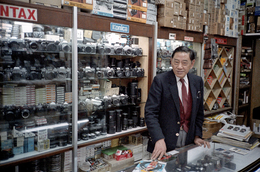 British Hong Kong, China - 1983: A vintage 1980's Fujifilm negative film scan of a camera store salesman in front of shelves of cameras and lenses for sale.