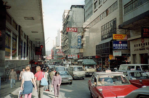British Hong Kong, China - 1983: A vintage 1980's Fujifilm negative film scan of busy city streets covered in store signs in Hong Kong with pedestrians, cars, and shops.