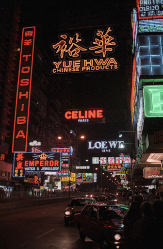 British Hong Kong, China - 1983: A vintage 1980's Fujifilm negative film scan of the downtown shopping centers of Hong Kong at night, with pedestrians, traffic, and illuminated shop signs.
