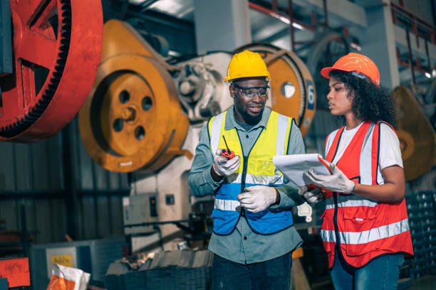istock Two professional engineer/worker/technician use clipboard discuss work, walk in steel metal manufacture factory plant industry. Black African American man and woman wear hard hat check quality machine 1417386458