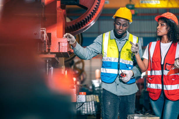 istock Two professional engineer/worker/technician use clipboard discuss work, walk in steel metal manufacture factory plant industry. Black African American man and woman wear hard hat check quality machine 1417386653