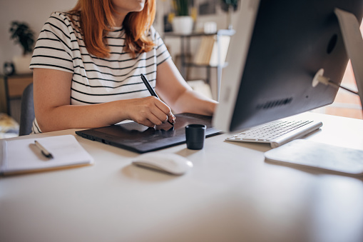 One woman, beautiful female sitting in his home office, using computer and working on graphics tablet for online course in graphics design.