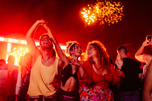 A group of young excited people are dancing in the night on a summer festival with fireworks in the background.