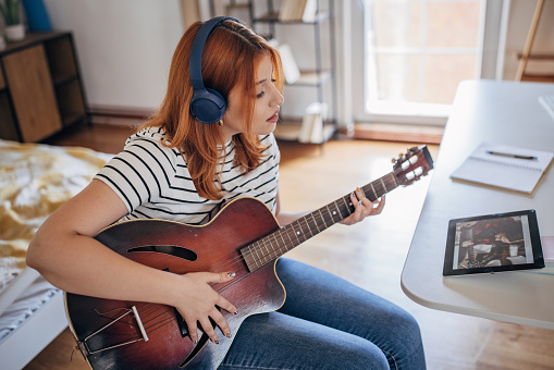 One woman, female with headphones sitting in bedroom at home, she is playing acoustic guitar while using tablet for online course.