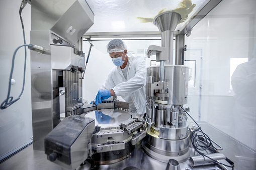 Pharmacy male technician wearing protective gloves, mask, cap and suit in a laboratory seen working on a specific machine that is the part of the pharmaceutical production during the working hours in a pharmaceutical factory.