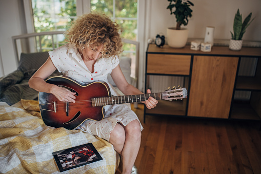 One woman, mature female with headphones sitting on bed in bedroom at home, she is playing acoustic guitar while using tablet for online course.