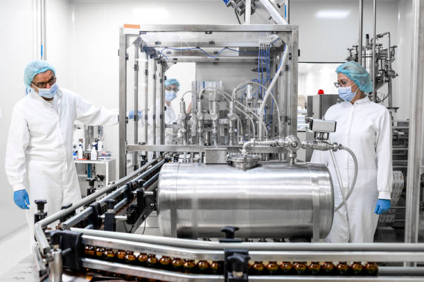 Two fully equipped coworkers in protective workwear seen in a pharmaceutical laboratory Two employees in pharmaceutical industry wearing protective gloves, mask, cap and white suit seen standing by the machine that is the part of the medicaments production during the working hours in a pharmaceutical manufacturing. drug manufacturing stock pictures, royalty-free photos & images