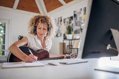 One woman, mature female with headphones sitting at the desk in bedroom at home, she is holding acoustic guitar while writing notes for online course.