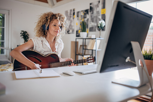 One woman, mature female with headphones sitting at the desk in bedroom at home, she is playing acoustic guitar while using computer for online course.