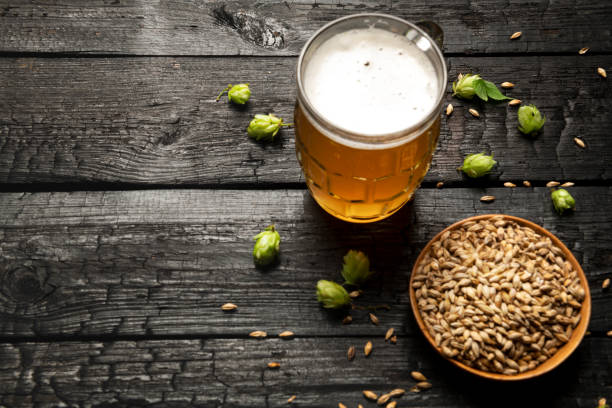 mug of light beer with foam and ingredients on a wooden table - rustic beer brewery indoors imagens e fotografias de stock