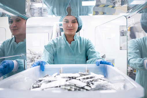 Young smiling female employee in a pharmaceutical laboratory completely dressed in a protective workwear seen holding a big plastic box with capsule pills in blister pack just after manufacturing process in a laboratory with mirror reflection.