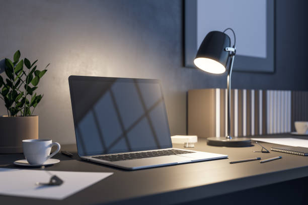 istock Side view on blank dark laptop screen with copyspace for your text or logo on dark wooden table with tea cup, stylish illuminated lamp and flower pot in the evening office. 3D rendering, mockup 1417397254