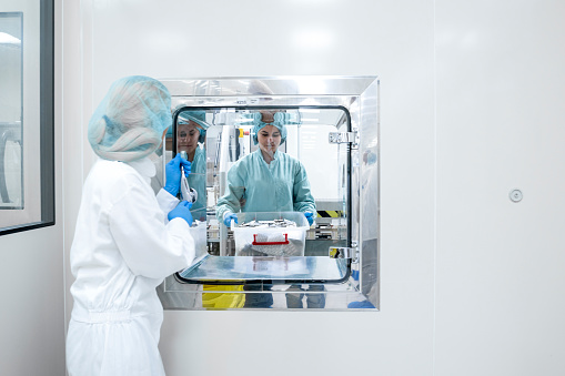 Two coworkers in a pharmaceutical factory completely dressed in a protective workwear seen while one is taking a big plastic box with capsule pills in blister pack from the other just after manufacturing process while her coworker is standing behind a glass window.