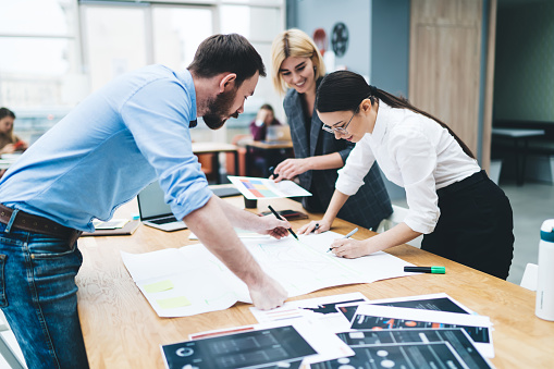 Smiling adult employees focusing and drawing blueprint while colleague assisting at big wooden desk with reports and laptop in office during daytime on blurred background