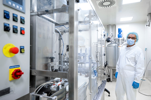 Male employee fully equipped in a protective workwear seen standing by the machine in a laboratory in a pharmaceutical factory.