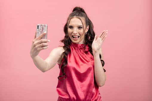 Portrait of a woman with long brown curly hair talking to a friend on the phone, video phone call, waving hand in greeting, pink background.