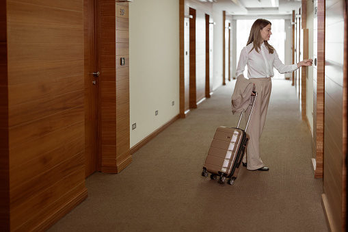 Elegant Business Lady in the Hallway of Hotel Room using Key Card while Entering her Room in a Luxury Hotel With Travel Trolley Luggage. Female Executive With Suitcase in Work Related Business Trip
