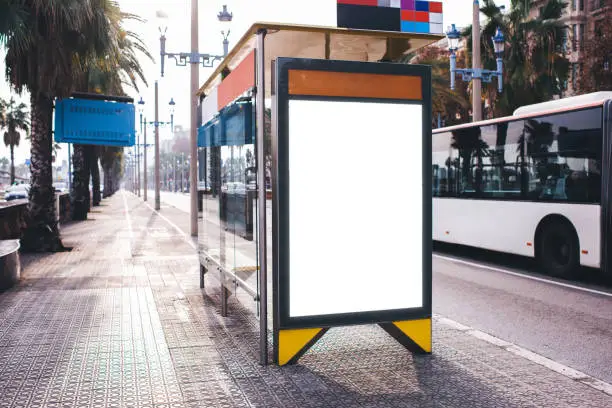 Public information board with blank screen for promotional message on bus stop located in urban setting with highway on sunny day