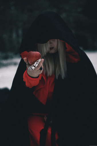 A woman in a witch costume for halloween in a winter forest offers a red apple in a black hood and red dress. A hand with rings holds out a red apple. Halloween concept, fairy tale.