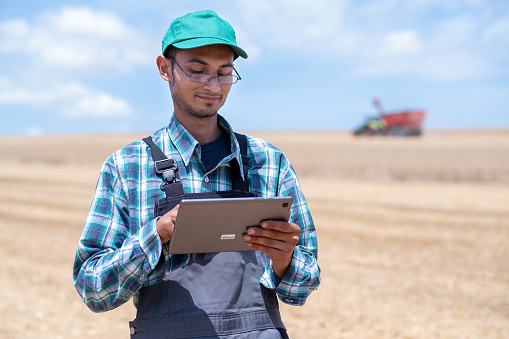 Young entrepreneur in gray overalls, green checkered shirt, green hat on wheat field during harvest.