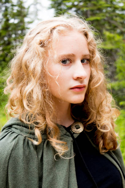 Teen or young adult girl wearing a green cloak looking at the camera against a lush forest background. Soft fantasy look