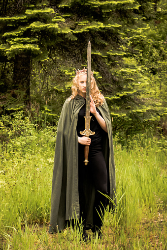 Teen or young adult girl wearing a crown and green cloak holding a long sword and looking at the camera against a lush forest background. Soft fantasy look